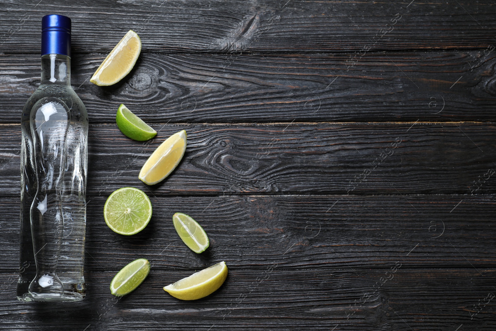 Photo of Bottle of vodka and citrus fruits on black wooden table, flat lay. Space for text