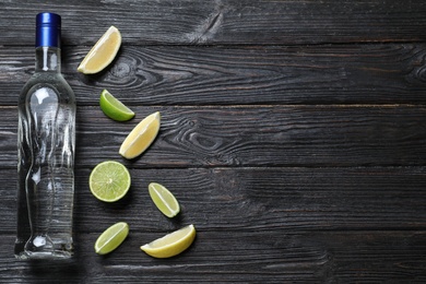 Bottle of vodka and citrus fruits on black wooden table, flat lay. Space for text