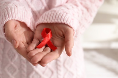 Photo of Woman holding red awareness ribbon indoors, closeup with space for text. World AIDS disease day