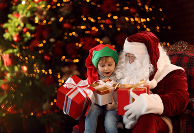 Photo of Santa Claus and little boy with gifts near Christmas tree indoors
