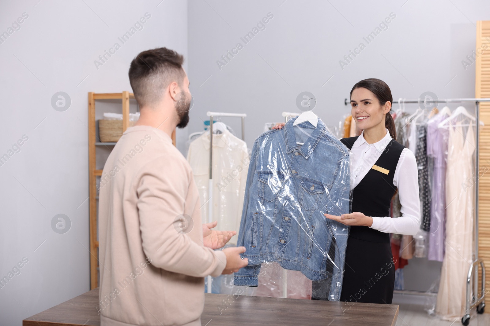 Photo of Dry-cleaning service. Happy worker showing denim jacket in plastic bag to client indoors