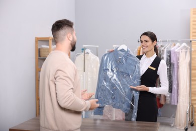 Dry-cleaning service. Happy worker showing denim jacket in plastic bag to client indoors