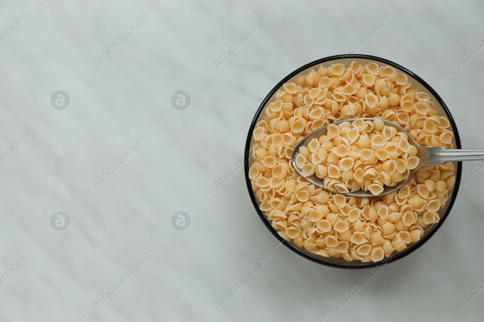 Photo of Bowl of uncooked conchiglie pasta and spoon on white marble table, top view. Space for text