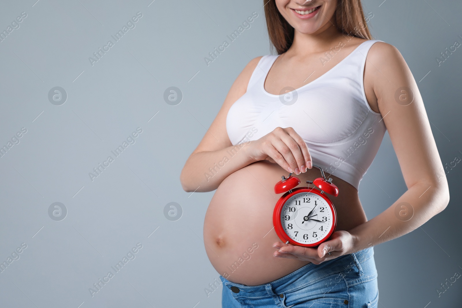 Photo of Young pregnant woman holding alarm clock near her belly on grey background, closeup and space for text. Time to give birth