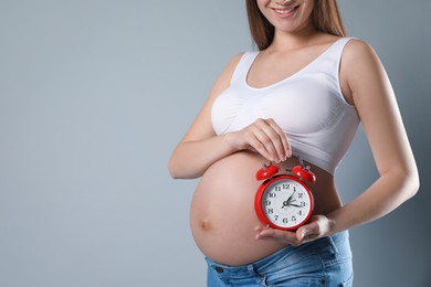 Photo of Young pregnant woman holding alarm clock near her belly on grey background, closeup and space for text. Time to give birth