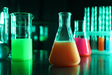 Photo of Laboratory glassware with luminous liquids on table against dark background