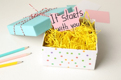 Photo of Open gift box and sheet of paper with words It Starts With You on white table