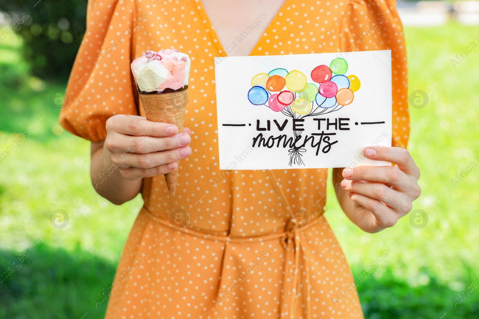 Photo of Woman holding card with phrase Live The Moments and ice cream outdoors, closeup