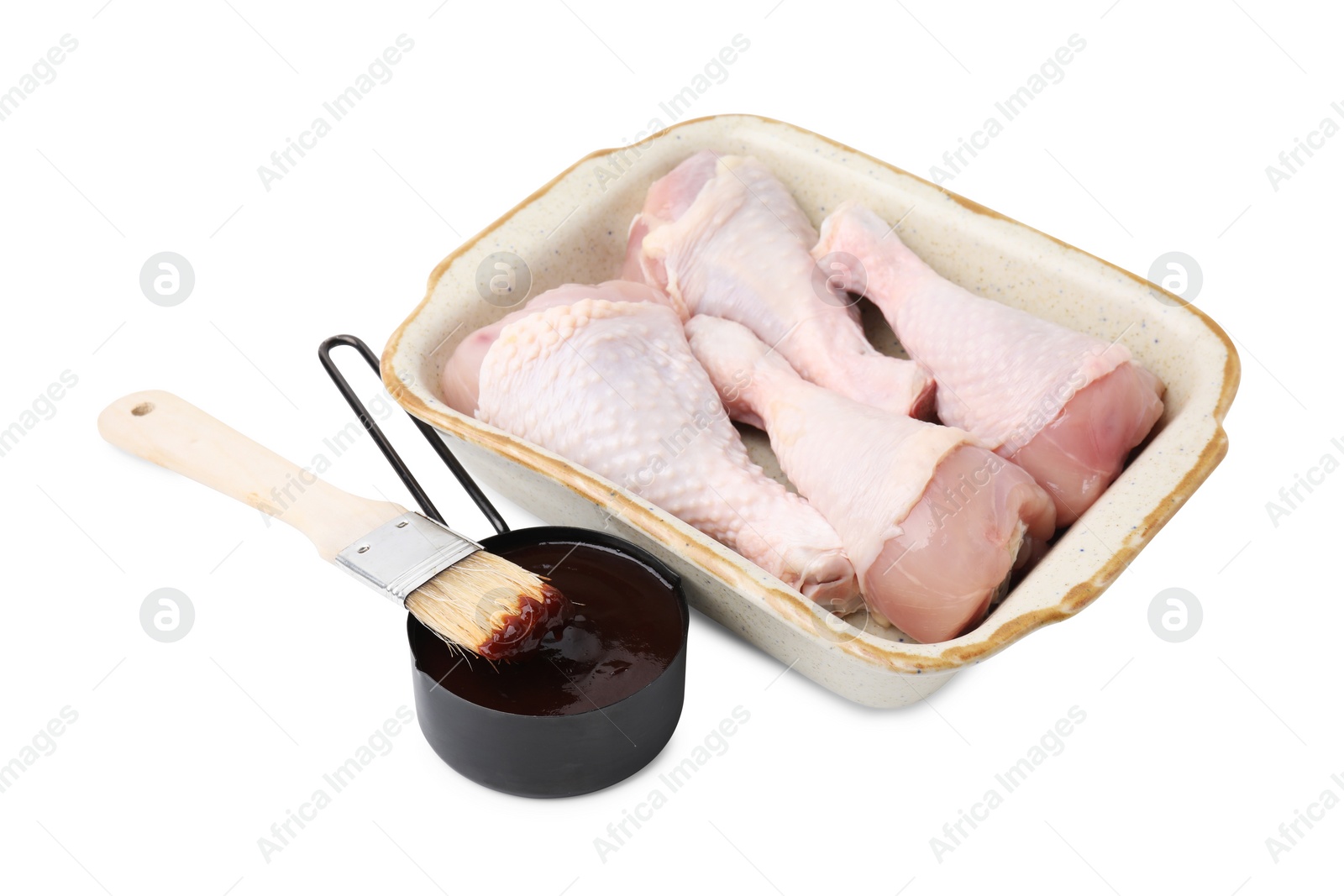 Photo of Raw chicken drumsticks in baking dish, marinade and basting brush isolated on white