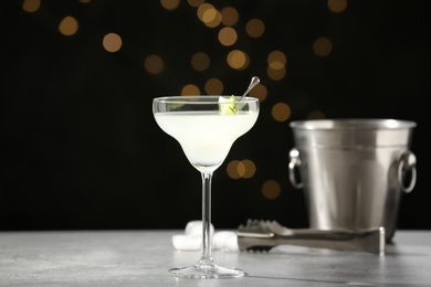 Photo of Glass of martini with cucumber, ice bucket and tongs on grey table against blurred lights. Space for text