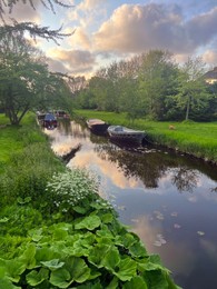 Canal with moored boats near green shore. Sky reflecting in river water