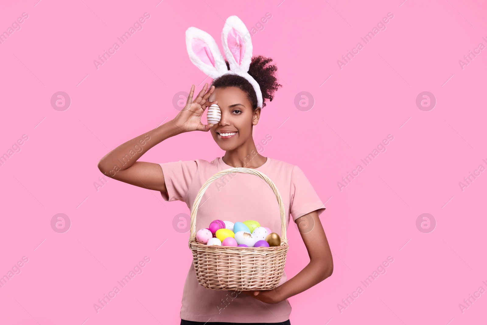 Photo of Happy African American woman in bunny ears headband covering eye with Easter egg on pink background