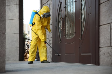 Photo of Person in hazmat suit disinfecting entrance door with sprayer. Surface treatment during coronavirus pandemic