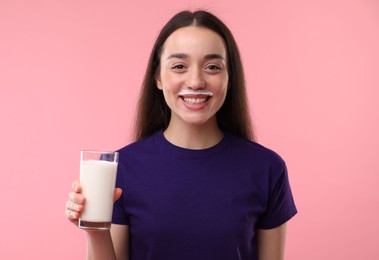 Smiling woman with milk mustache holding glass of tasty dairy drink on pink background