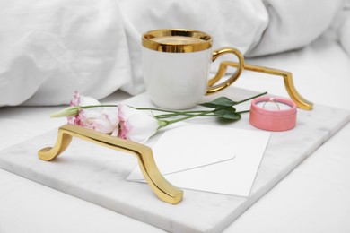 Photo of Tray with cup of coffee, flowers and beautiful engagement ring in box on white bed, closeup