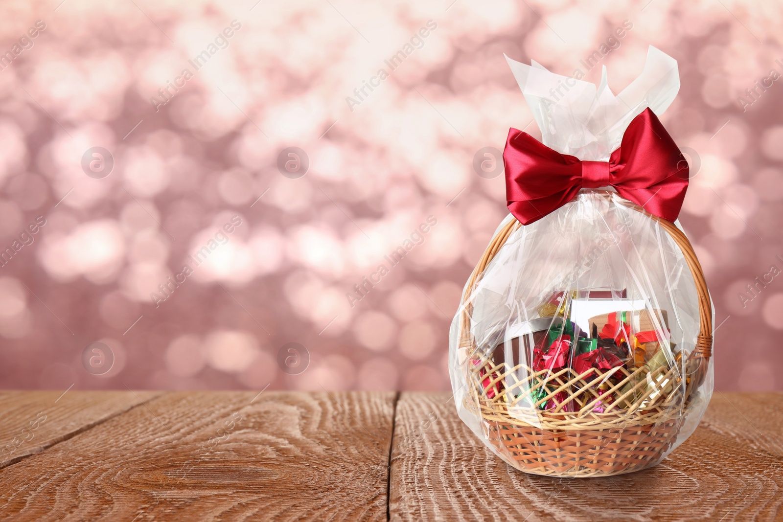 Image of Wicker basket full of gifts on wooden table against blurred festive lights. Space for text