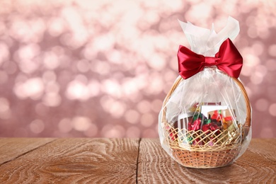 Image of Wicker basket full of gifts on wooden table against blurred festive lights. Space for text