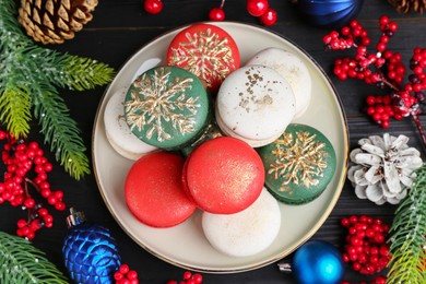 Different decorated Christmas macarons and festive decor on black wooden table, flat lay