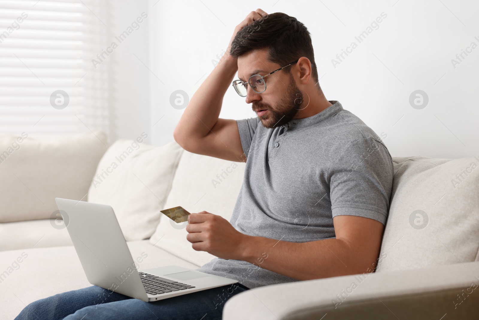 Photo of Upset man with credit card and laptop on sofa at home. Be careful - fraud