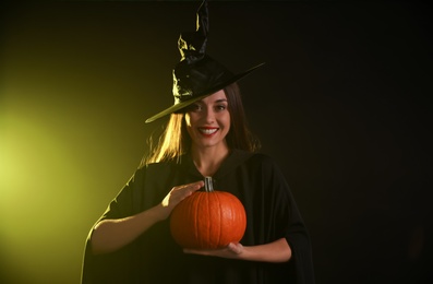 Photo of Young woman wearing witch costume with pumpkin on dark background. Halloween party