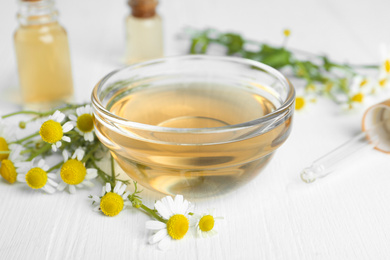 Bowl of essential oil and fresh chamomiles on white wooden table