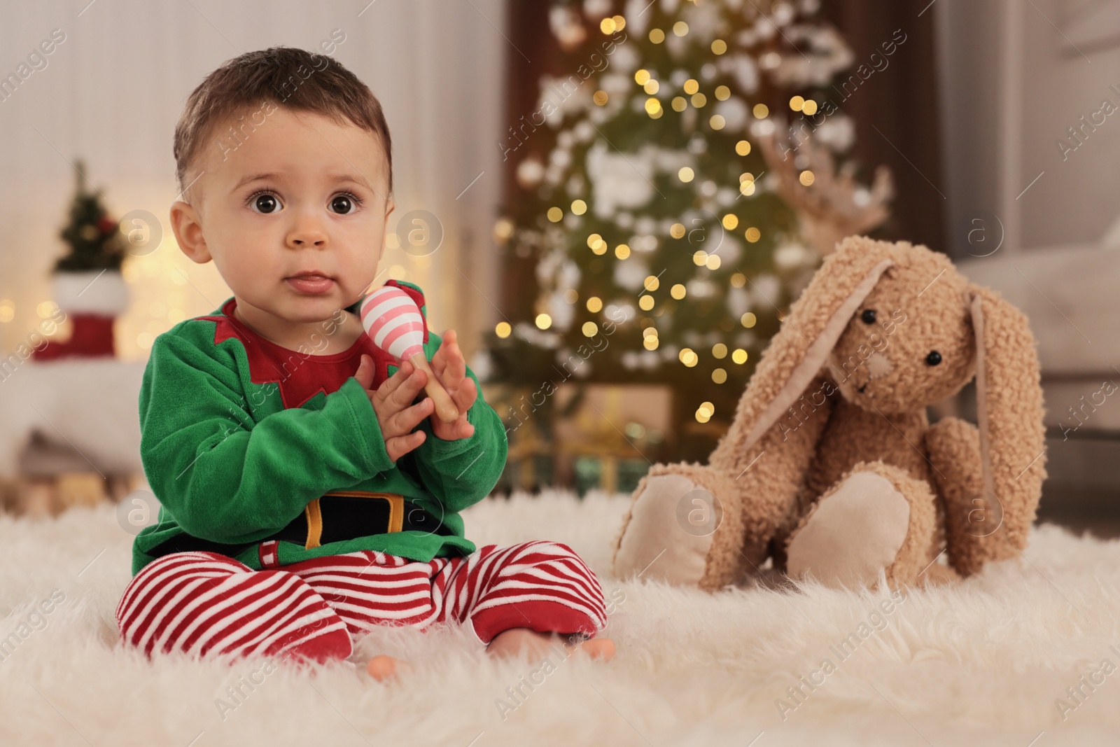 Photo of Baby wearing cute elf costume on floor in room decorated for Christmas