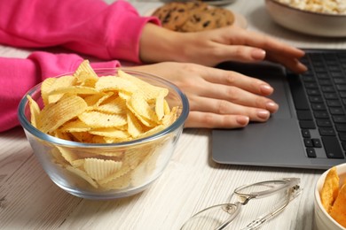 Photo of Bad eating habits. Woman working on laptop at white wooden table with ridged chips in bowl, selective focus