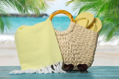 Image of Beach bag with towel, flip flops and heart shaped sunglasses on light blue wooden surface near seashore