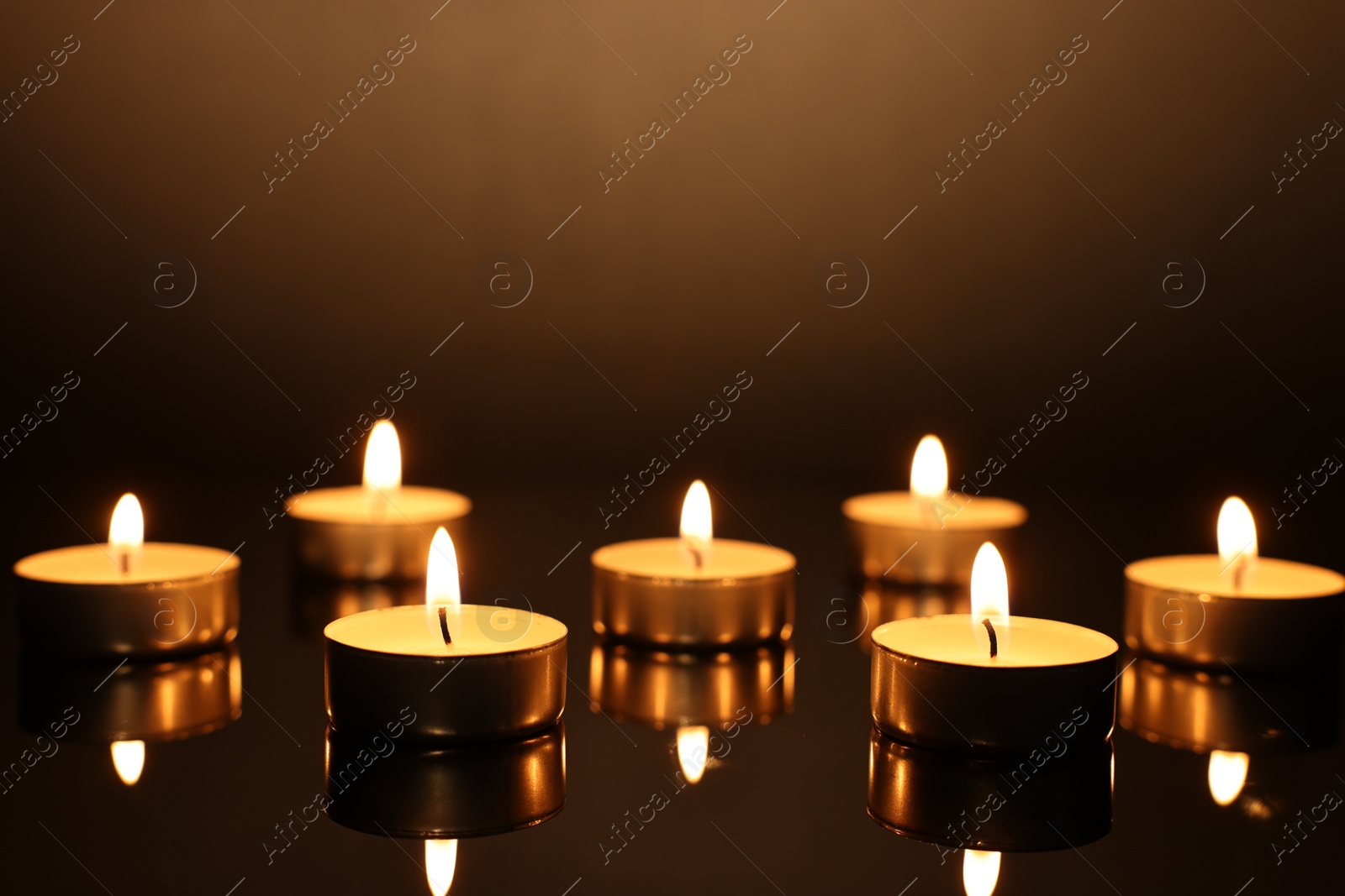 Photo of Burning candles on mirror surface in darkness, closeup
