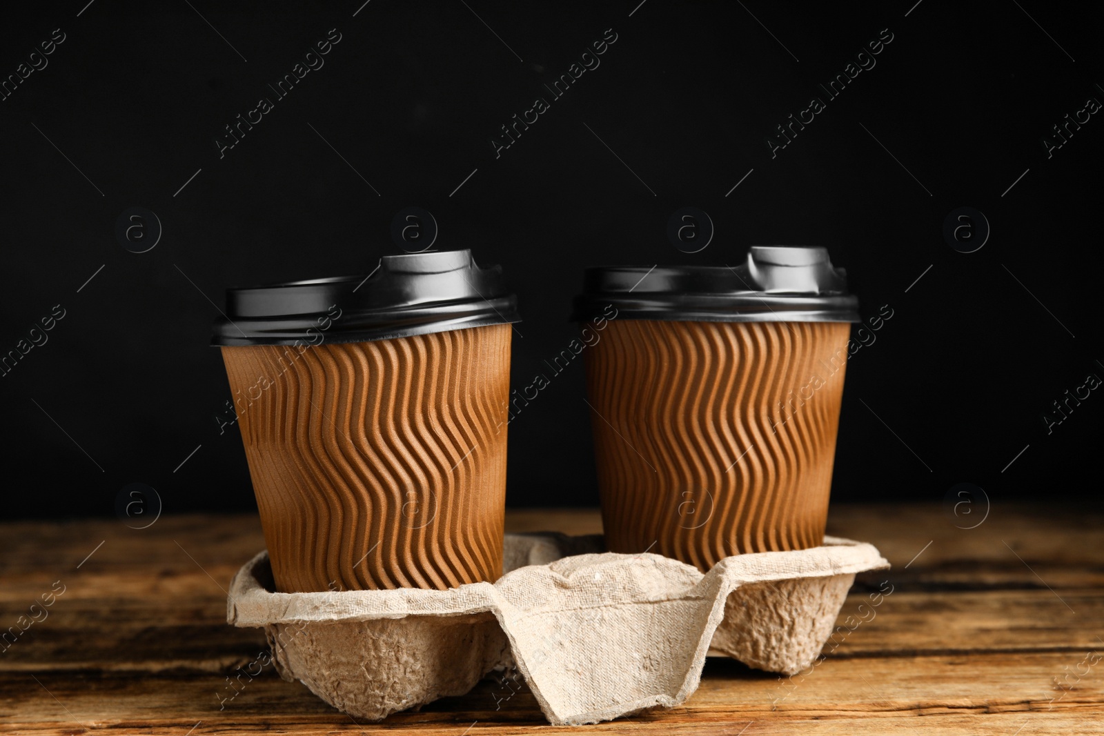 Photo of Takeaway paper coffee cups in cardboard holder on wooden table against black background