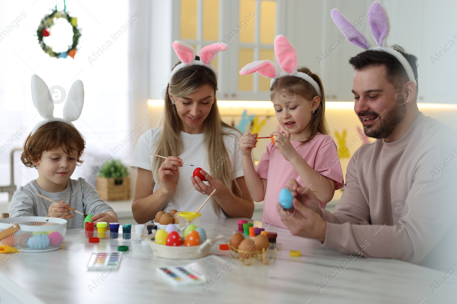 Photo of Easter celebration. Happy family with bunny ears painting eggs at white marble table in kitchen