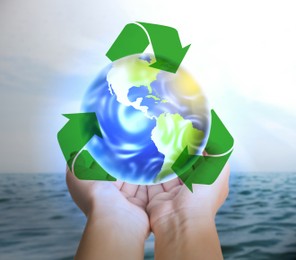 Image of Woman holding virtual image of Earth and recycling symbol near ocean, closeup view