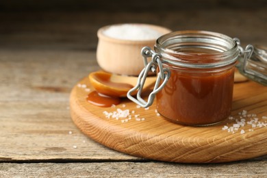 Photo of Tasty salted caramel in jar and salt on wooden table, closeup. Space for text