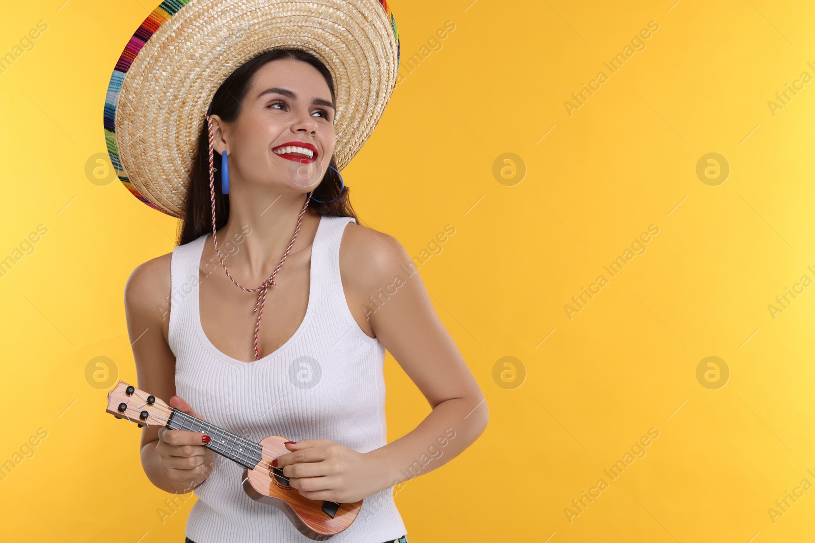 Photo of Young woman in Mexican sombrero hat playing ukulele on yellow background. Space for text