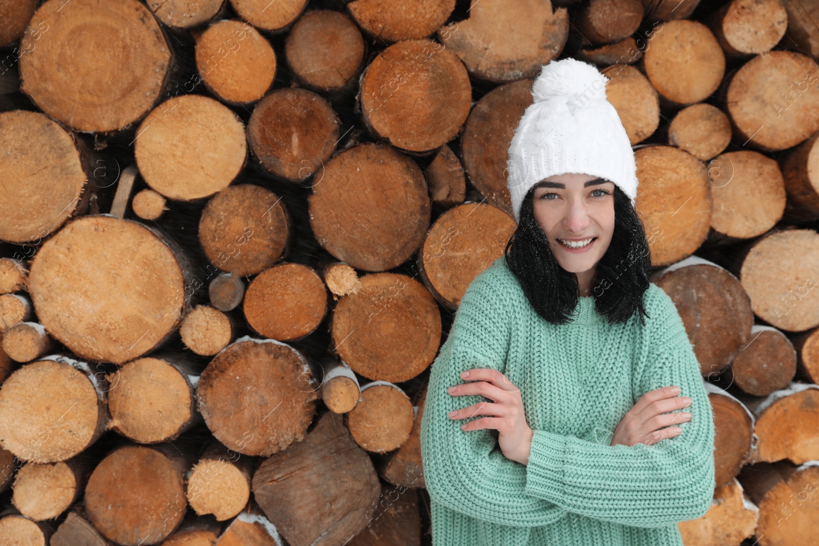 Photo of Young woman wearing warm sweater and hat near stack of firewood, space for text. Winter season