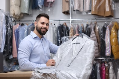 Photo of Dry-cleaning service. Happy worker holding hanger with jacket in plastic bag at counter indoors