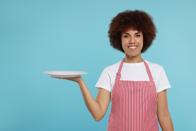 Happy young woman in apron holding plate on light blue background