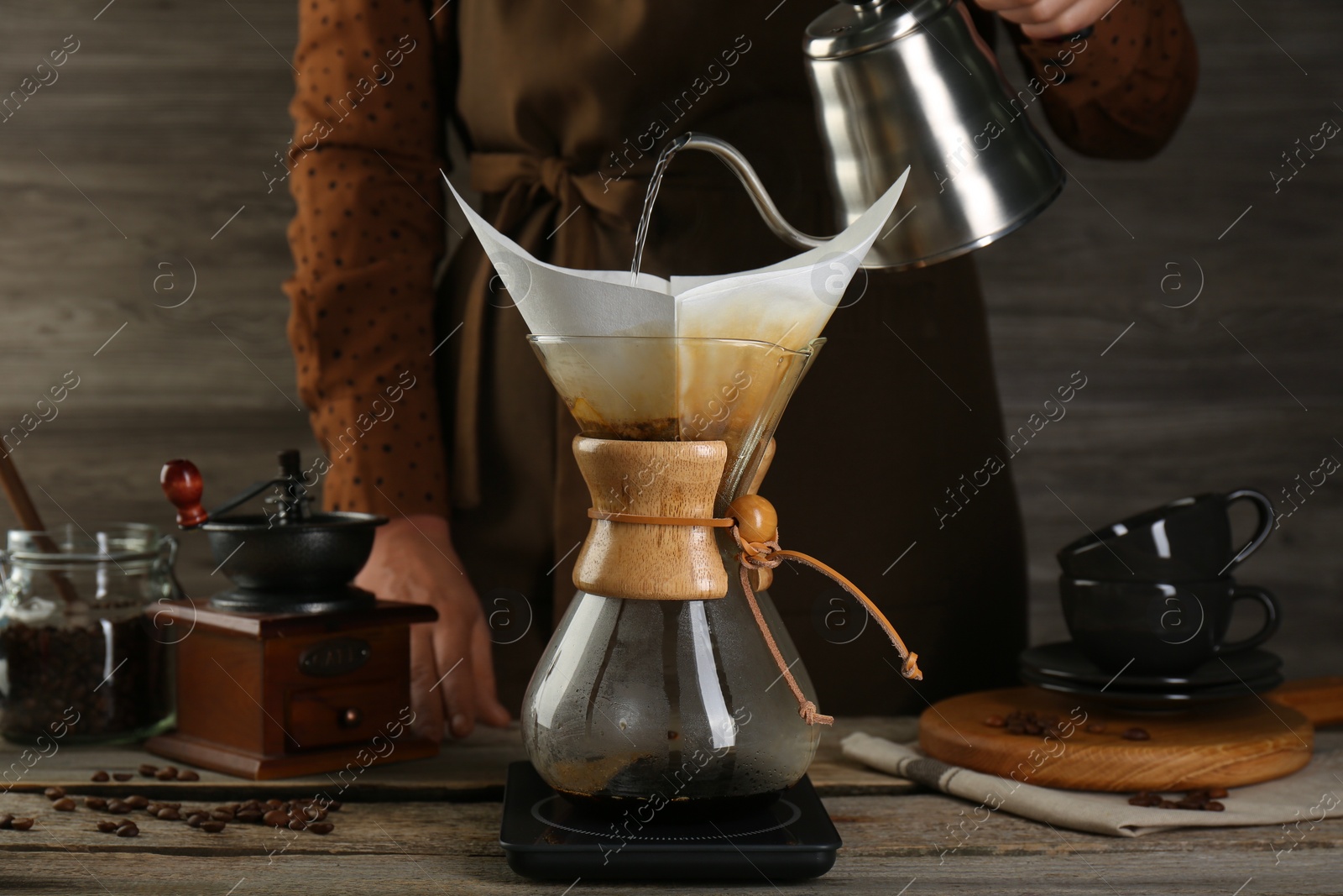 Photo of Woman pouring hot water into glass chemex coffeemaker with paper filter at wooden table, closeup