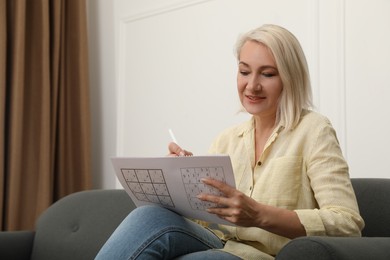 Photo of Middle aged woman solving sudoku puzzle on sofa at home