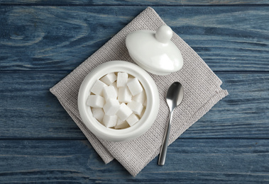 Photo of Refined sugar cubes in ceramic bowl on blue wooden table, top view