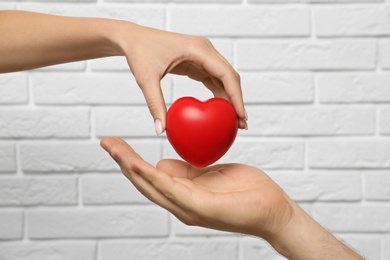 Woman giving red heart to man at white brick wall, closeup. Donation concept