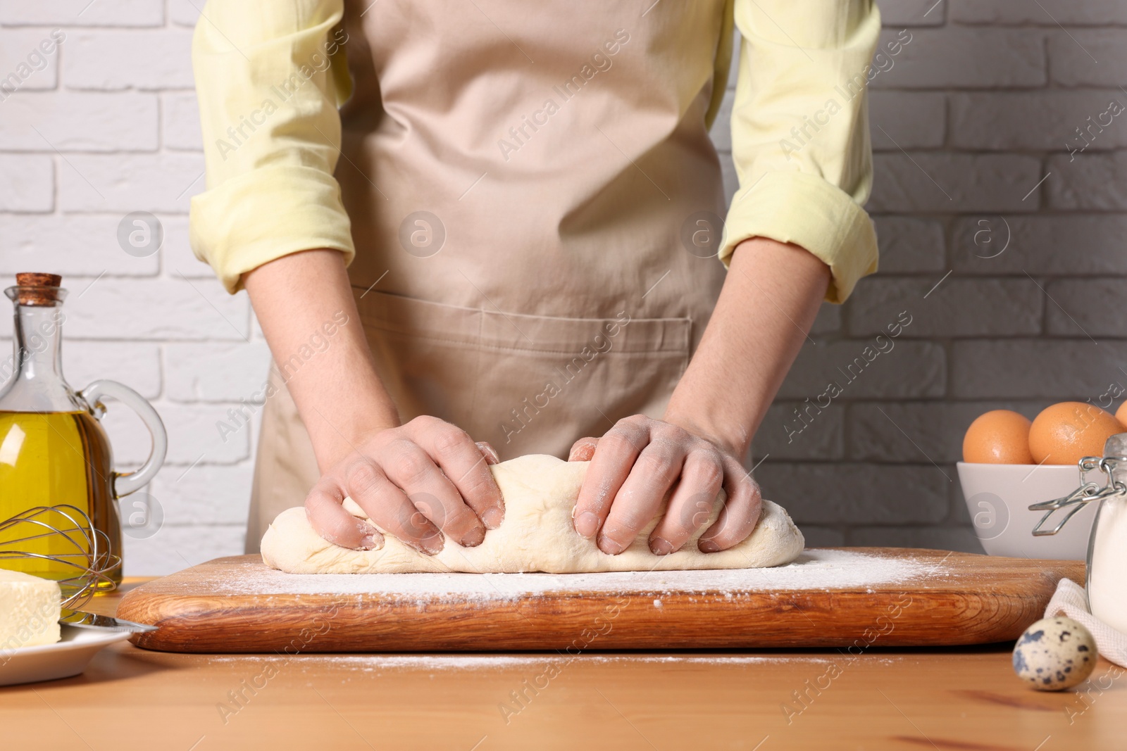 Photo of Woman kneading dough at wooden table near white brick wall, closeup