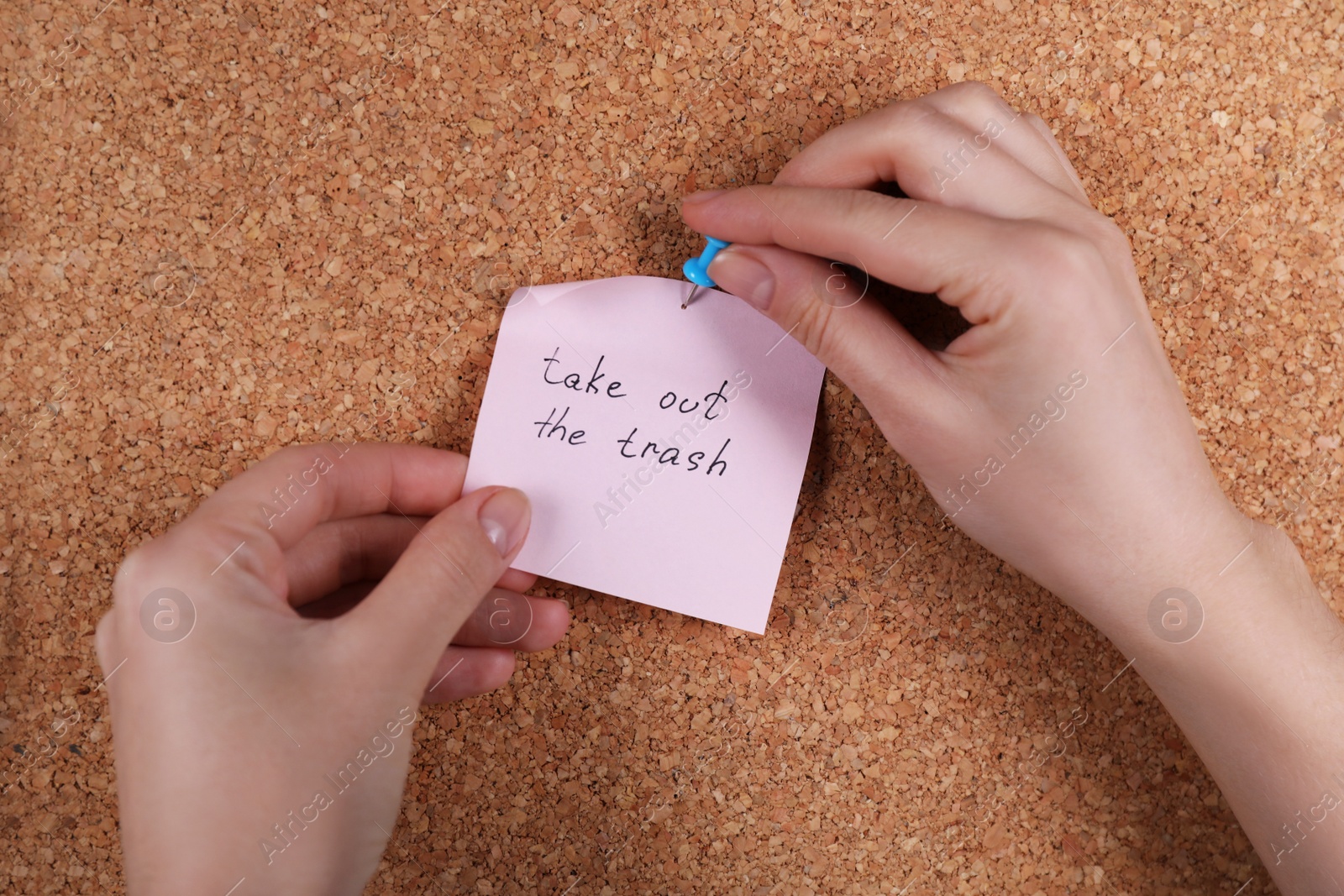 Photo of Woman pinning paper note with phrase Take Out The Trash to cork board, closeup