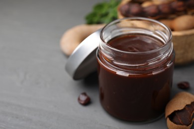 Photo of Jar of tasty tamarind sauce and fresh pods on grey wooden table. Space for text