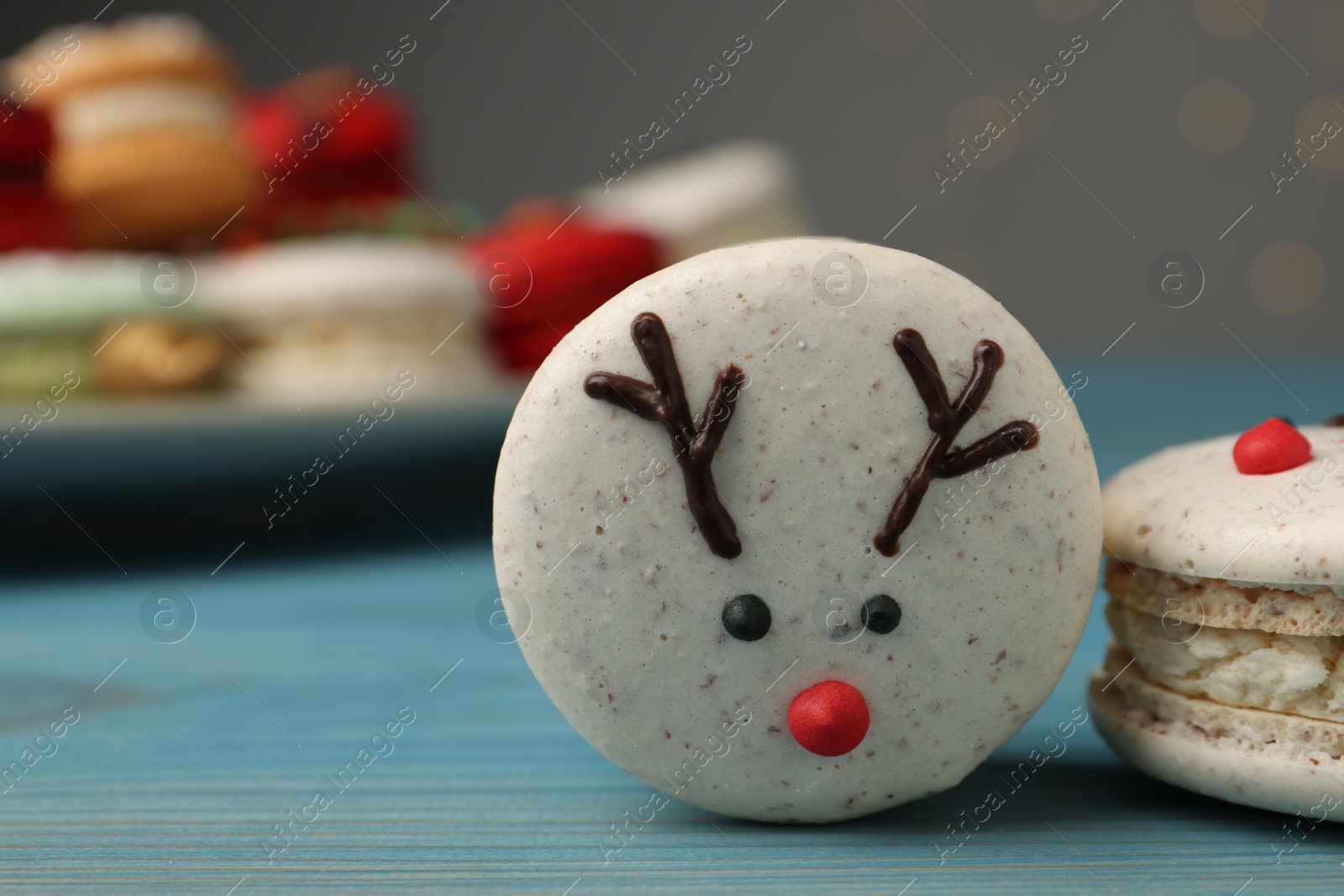 Photo of Tasty reindeer Christmas macarons on light blue wooden table against blurred festive lights, closeup