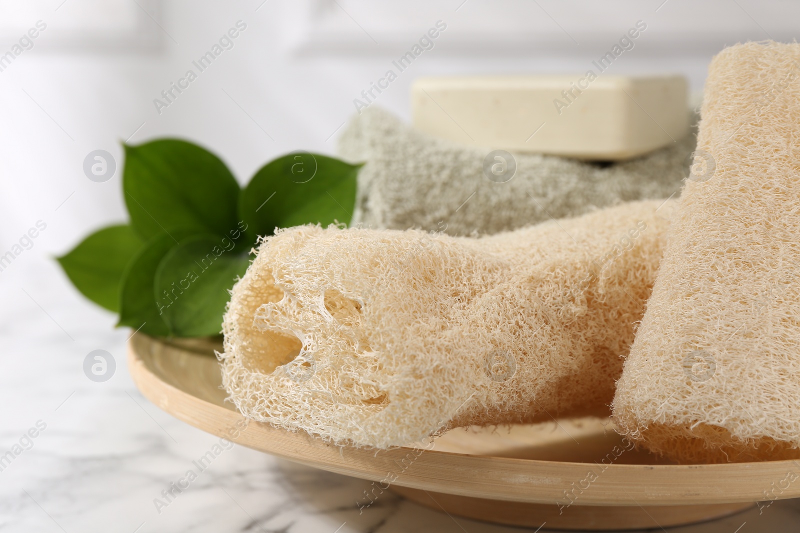Photo of Loofah sponges, soap, towel and green leaves on white marble table, closeup