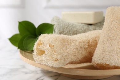 Loofah sponges, soap, towel and green leaves on white marble table, closeup