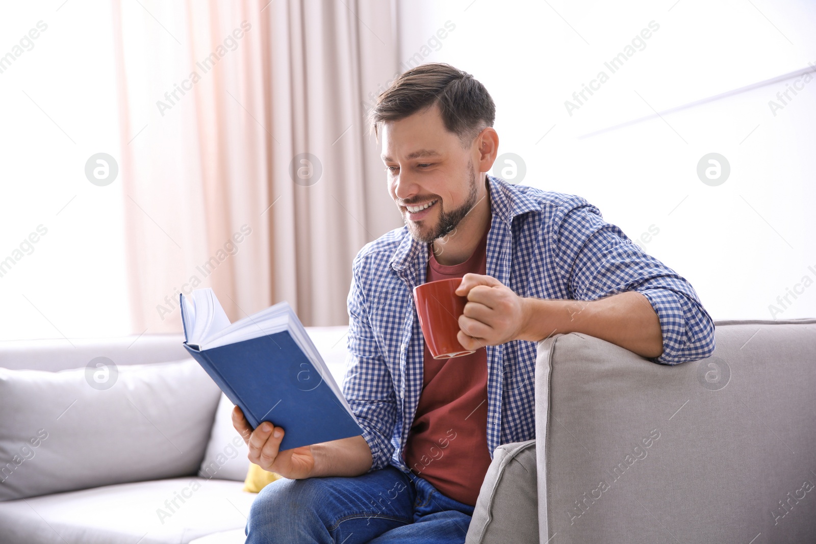 Photo of Handsome man with cup of coffee reading book on sofa at home