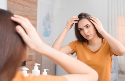 Photo of Young woman with hair loss problem looking in mirror indoors