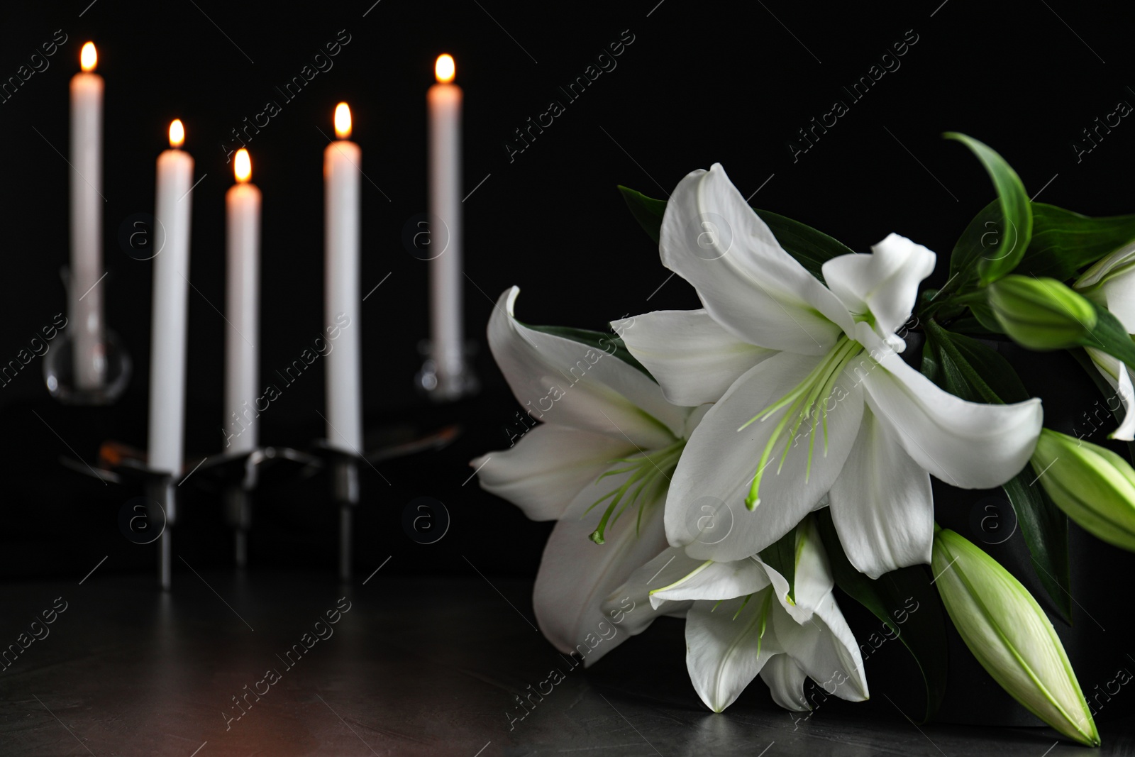 Photo of White lilies and blurred burning candles on table in darkness, closeup with space for text. Funeral symbol